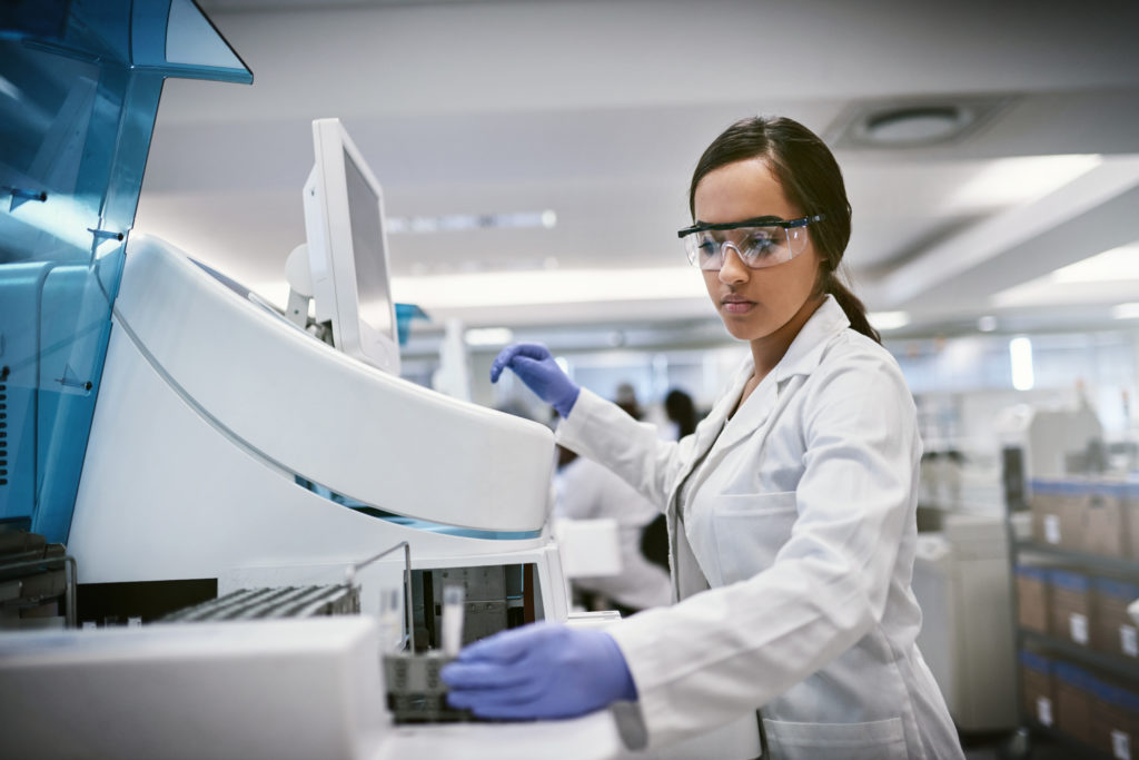 Woman in a lab coat and goggles in front of a machine