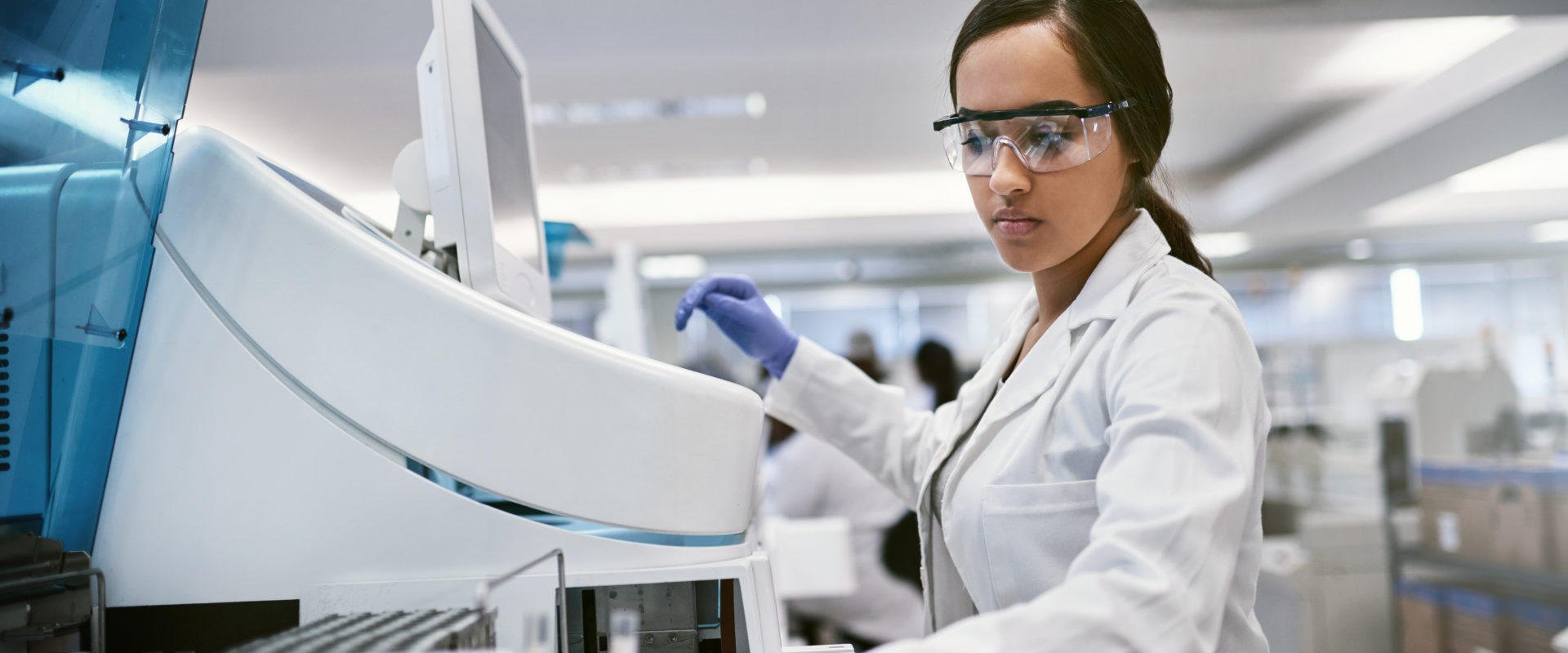 Woman in a lab coat and goggles in front of a machine
